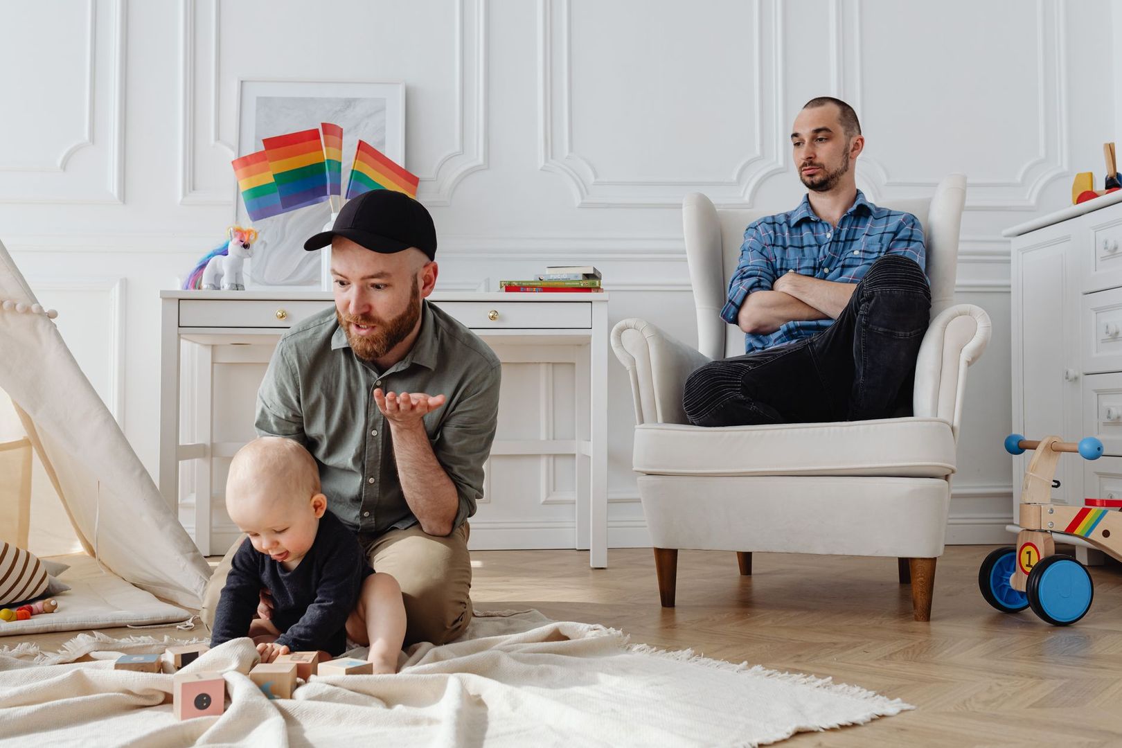 En pappa leker med sitt barn på golvet medan den andra pappan tittar på från en fåtölj.