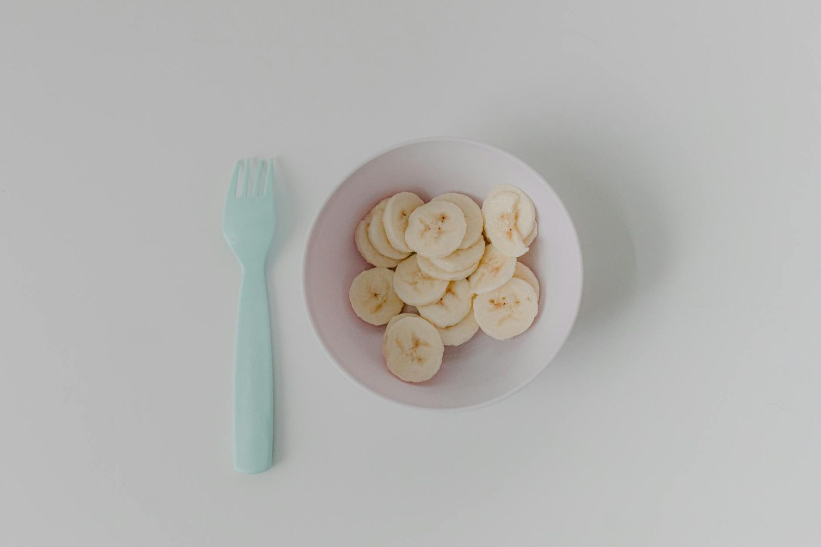 Skivade bananer i en rosa skål bredvid en ljusblå gaffel.